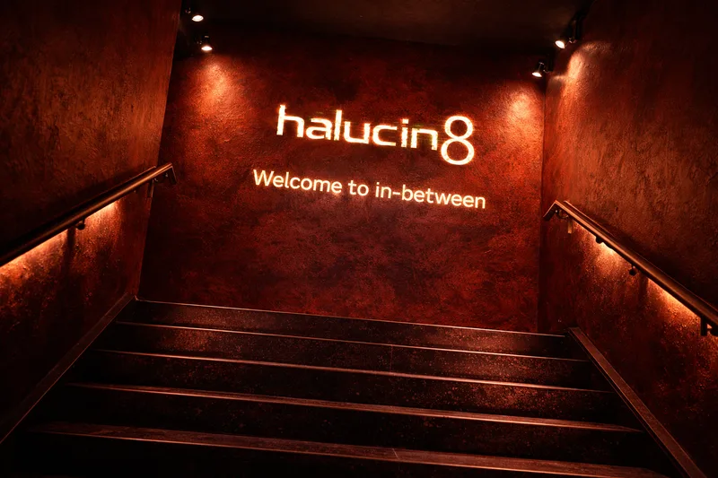Red-lit stairwell rising to a textured wall with the glowing halucin8 wordmark and the line Welcome to in-between